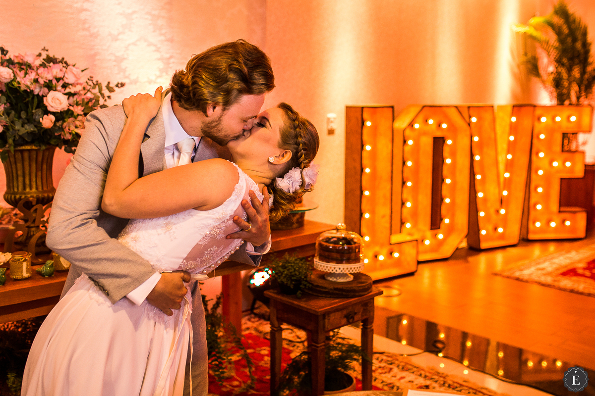 beijo dos noivos em cerimonia de casamento com decoração retro e diferente ao lado do love gigante de luzes