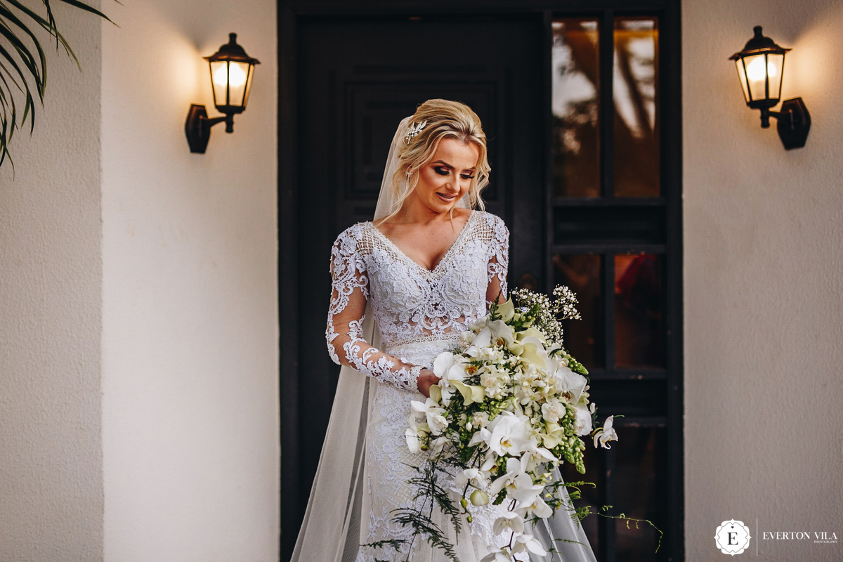 linda noiva com vestido de noiva classico de manga longa e renda segurando seu buque com orquideas brancas em frente ao estudio da le pequeno em maringa