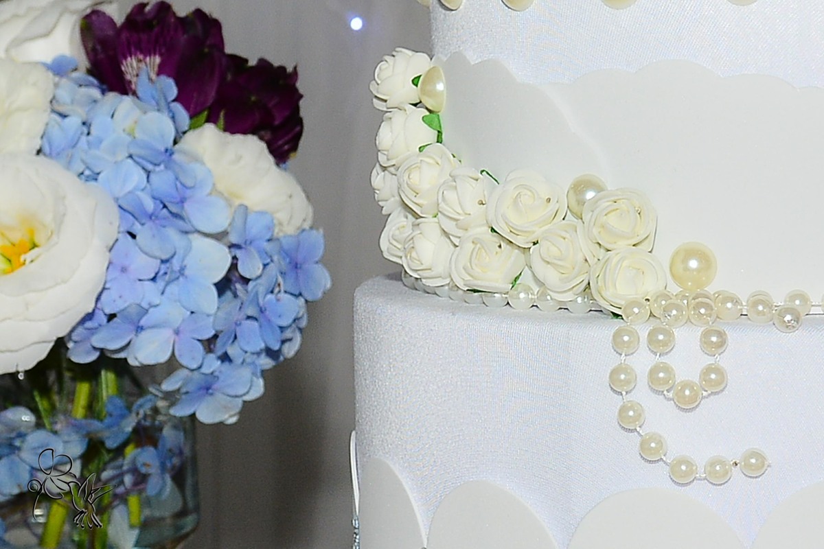 by Eli Matos detalha o bolo, aparecem rosas na cor perolada e pérolas, ao lado flores azuis.
