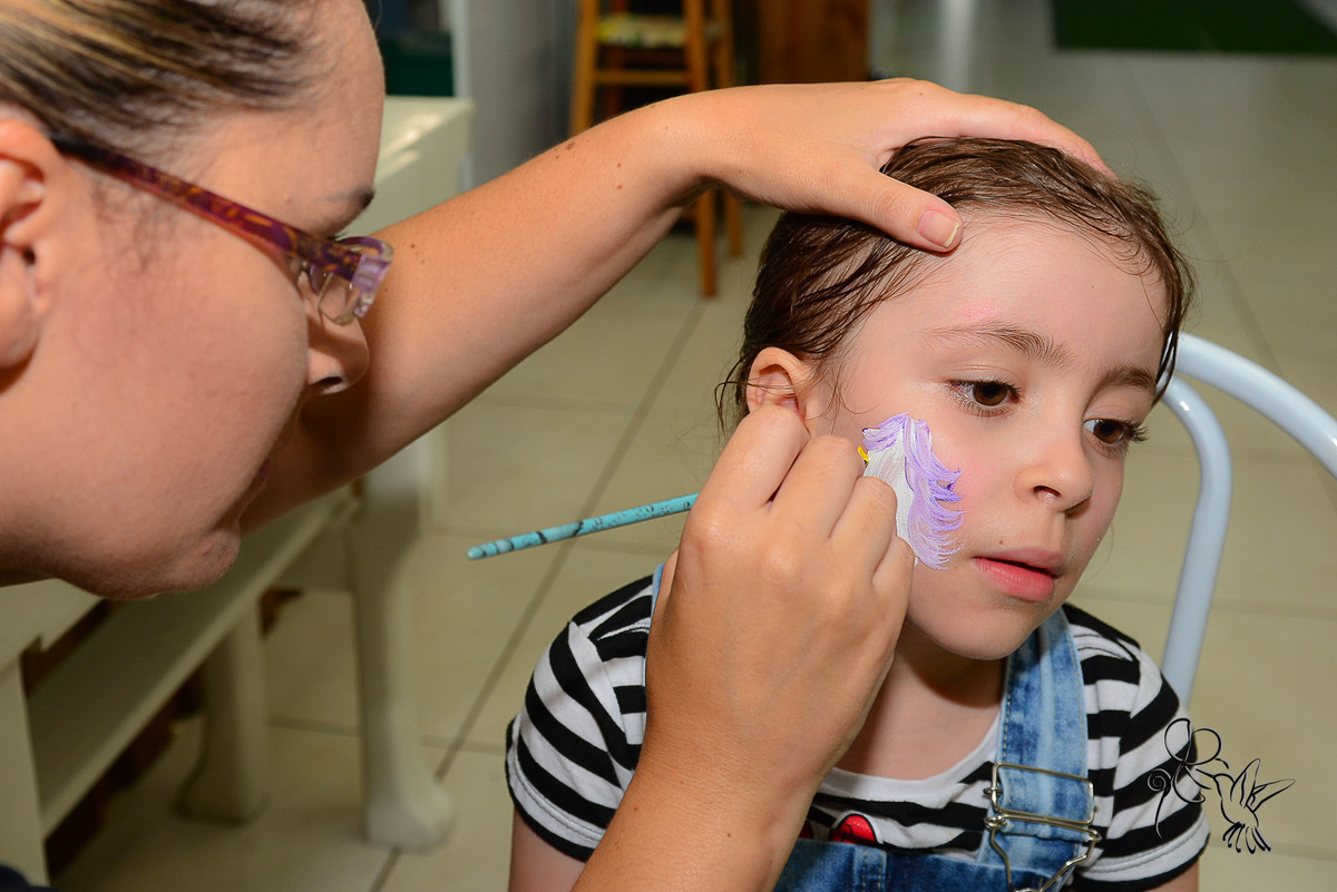 Pessoa faz pintura de rosto em tons de lilás em uma menina, Menino de 4 anos caminha feliz  no seu aniversário, clique de by Eli Matos quem registrou