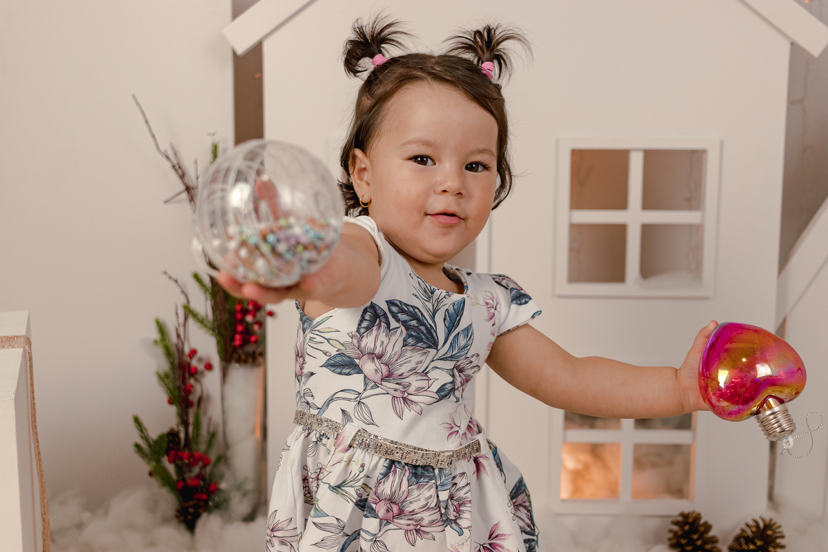 menina oriental em frente cenario de natal segurando uma bolinha colorida em uma das mãos e outra em formato de coração na outra mão