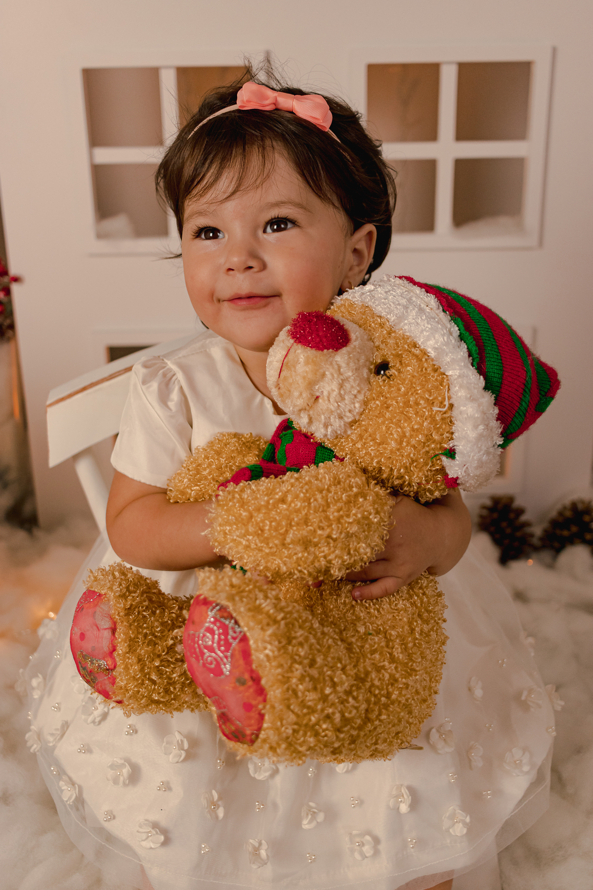 menina oriental segurando urso de pelucia com toca de papi noel sentada em uma cadeira em frente cenario natalino de um estudio