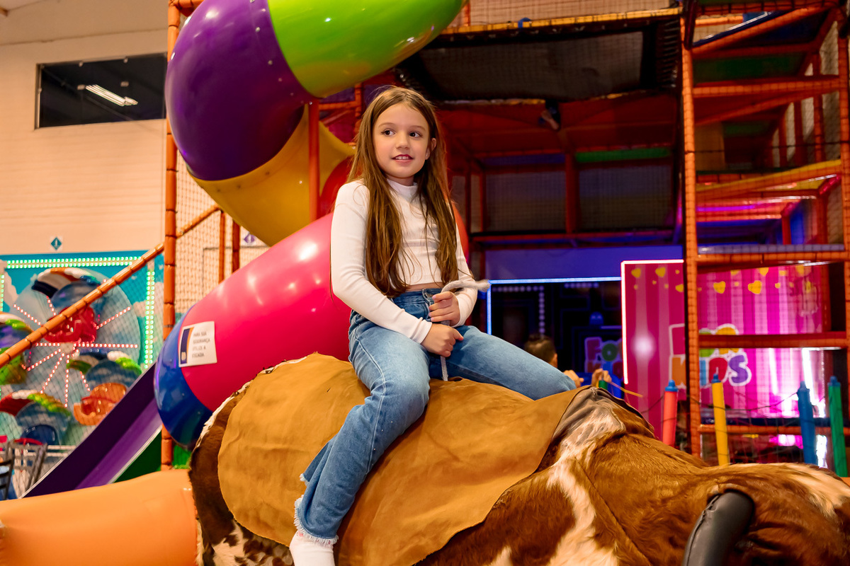 menina de calça jeans e camisa branca em cima de um touro mecânico em uma casa de festas