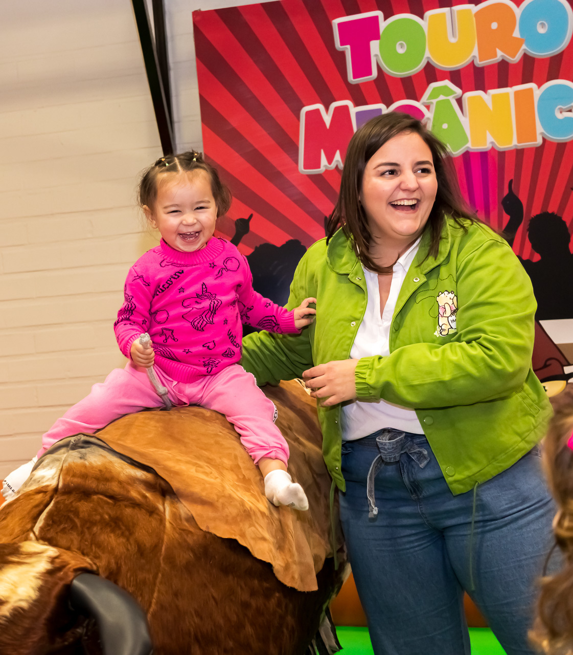 bebezinho em cima de touro mecânico sendo segurada por mulher em pé do lado do touro