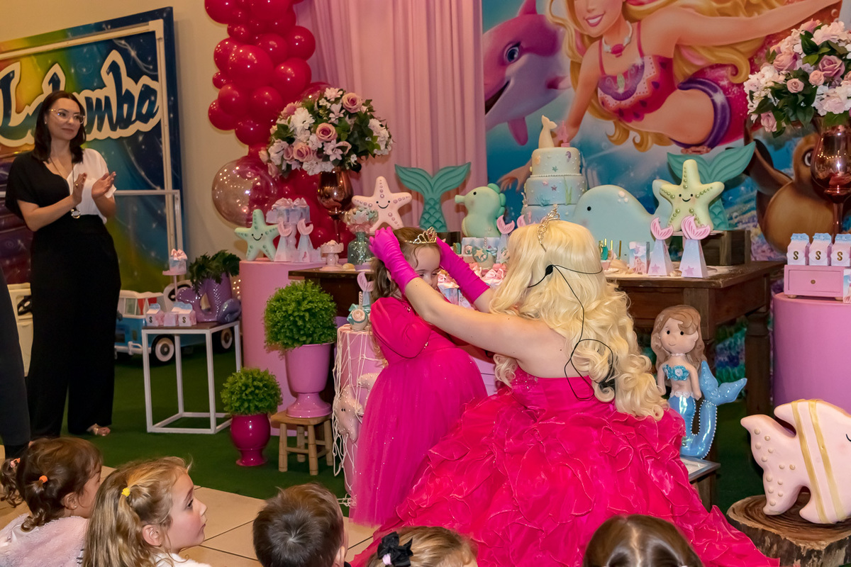 no fundo decoração da Barbie e na frente a Barbie princesa colocando coroa na anivesariante. Ambasa estão de vestido pink