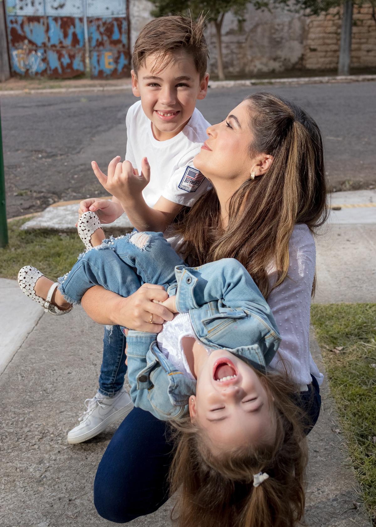 mãe abaixada segurando filha de cabeça para baixo e olhando para o filho que está atrás delas