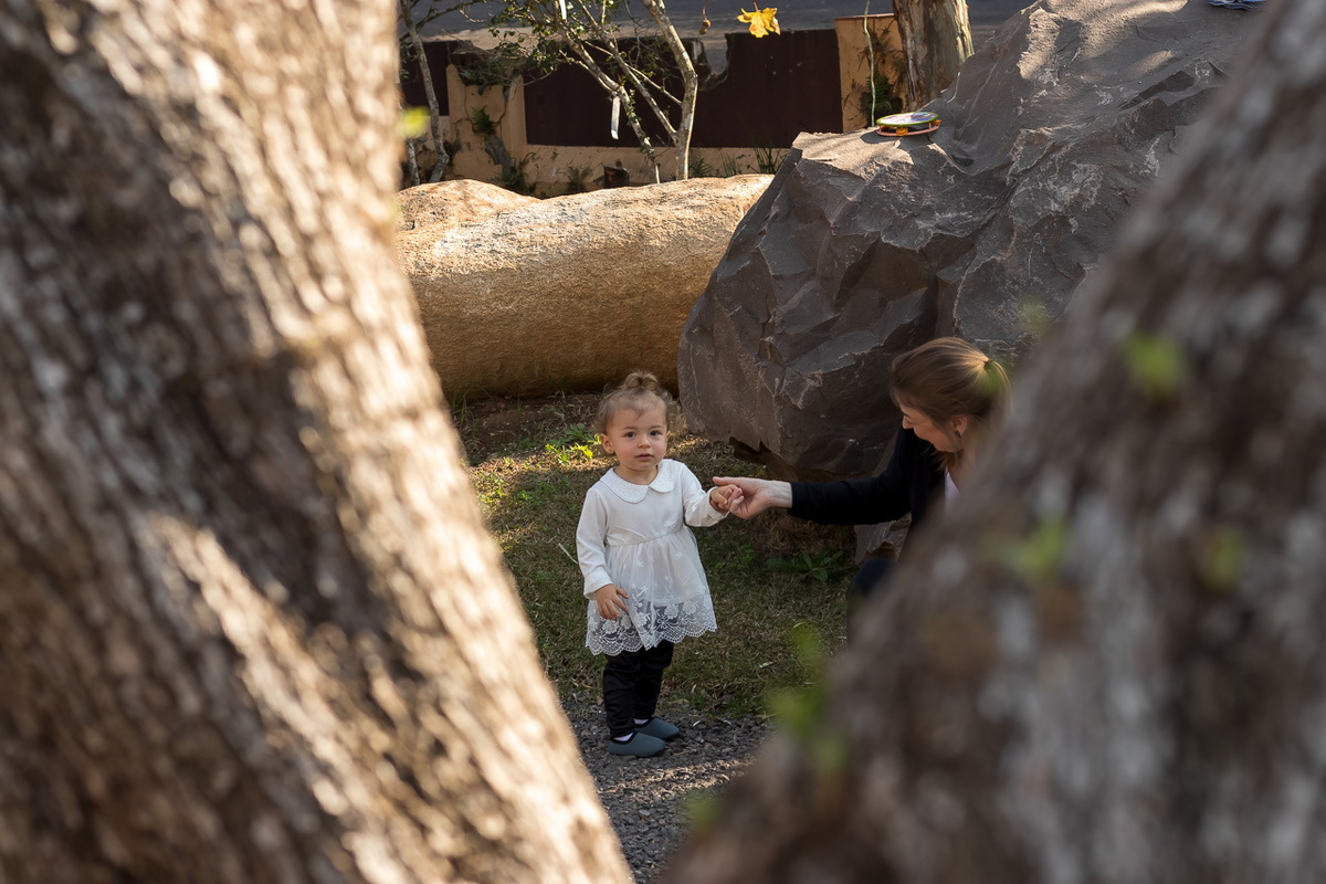 criança de vestido branco mais para o fundo e mais em primeiro plano dois troncos de arvore em V que fazem a moldura da criança 