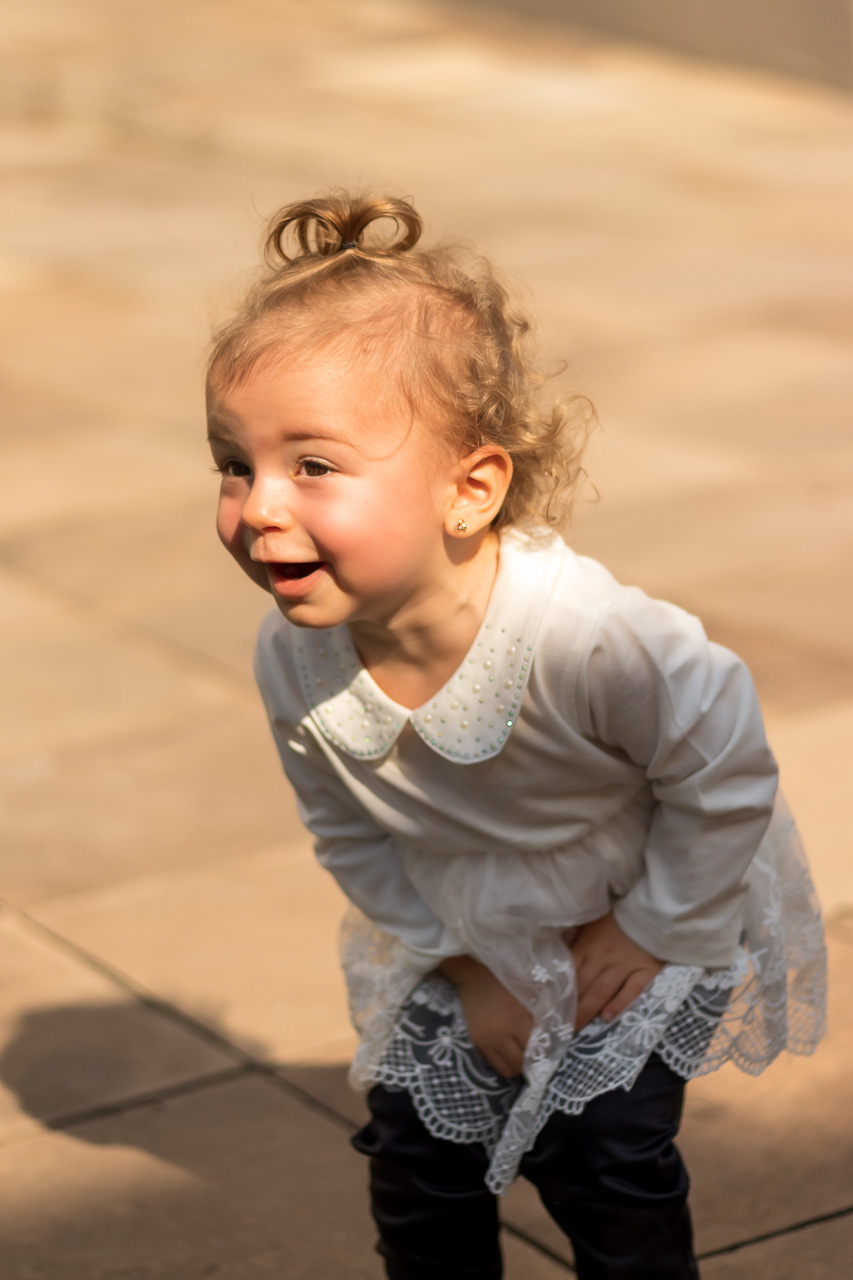 criança levemente inclinada pra frente com sorriso . de vestido branco . cabelos loirinhos e cacheados