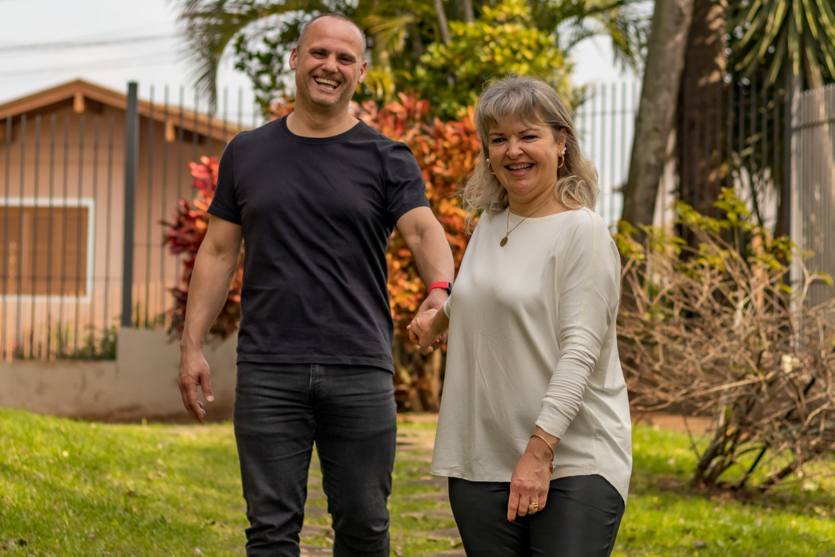 casal de mãos dadas olhado para câmera. Ela na frente puxando a mão dele. Foto tirada no pátio da casa.