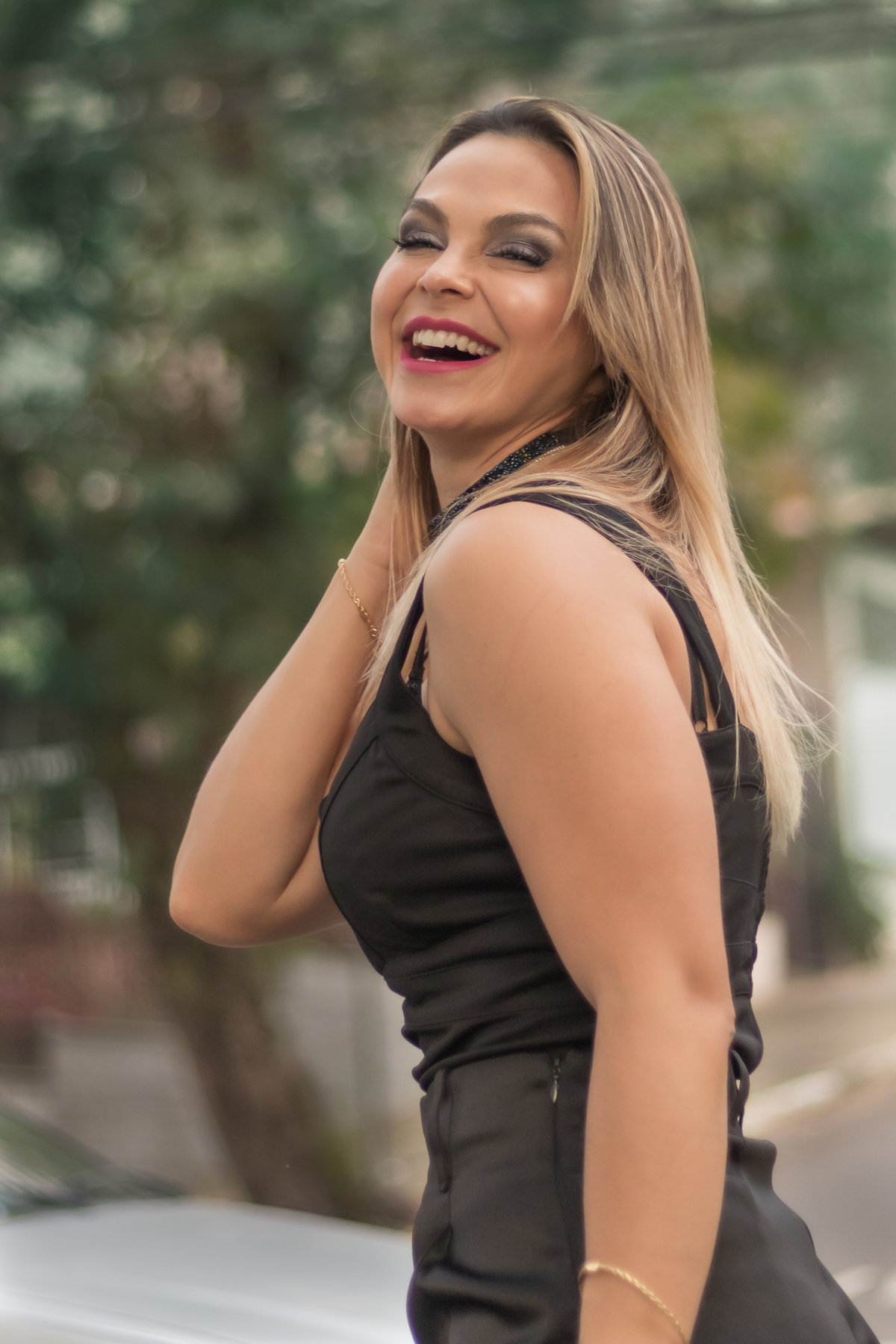 modelo de lado sorrindo com mão direta segurado pescoço e prendendo cabelo. vestido preto e fundo desfocado