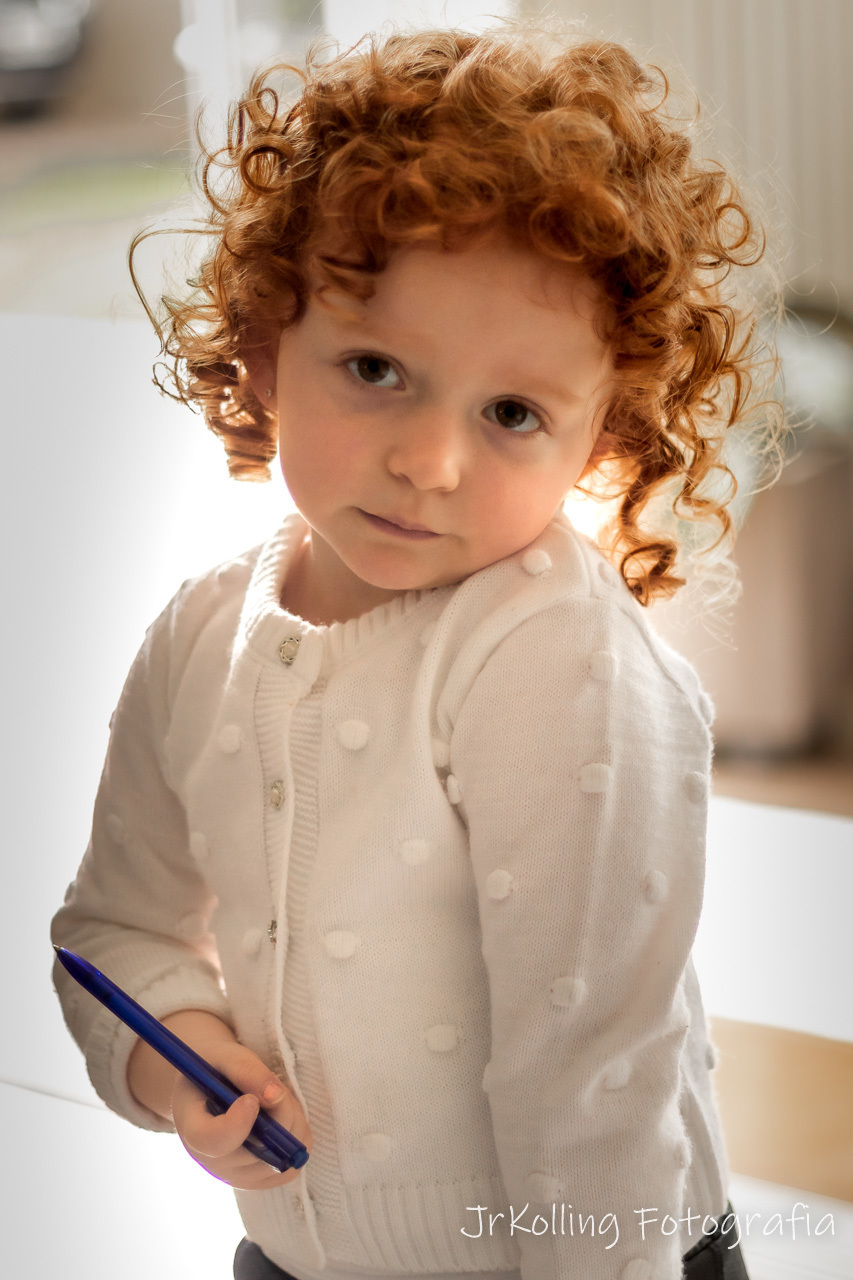 menininha ruiva olhando para a camera com pescoço levemente inclinado para esquerda encostando no ombro. casaco branco segurando uma caneta com mão direita