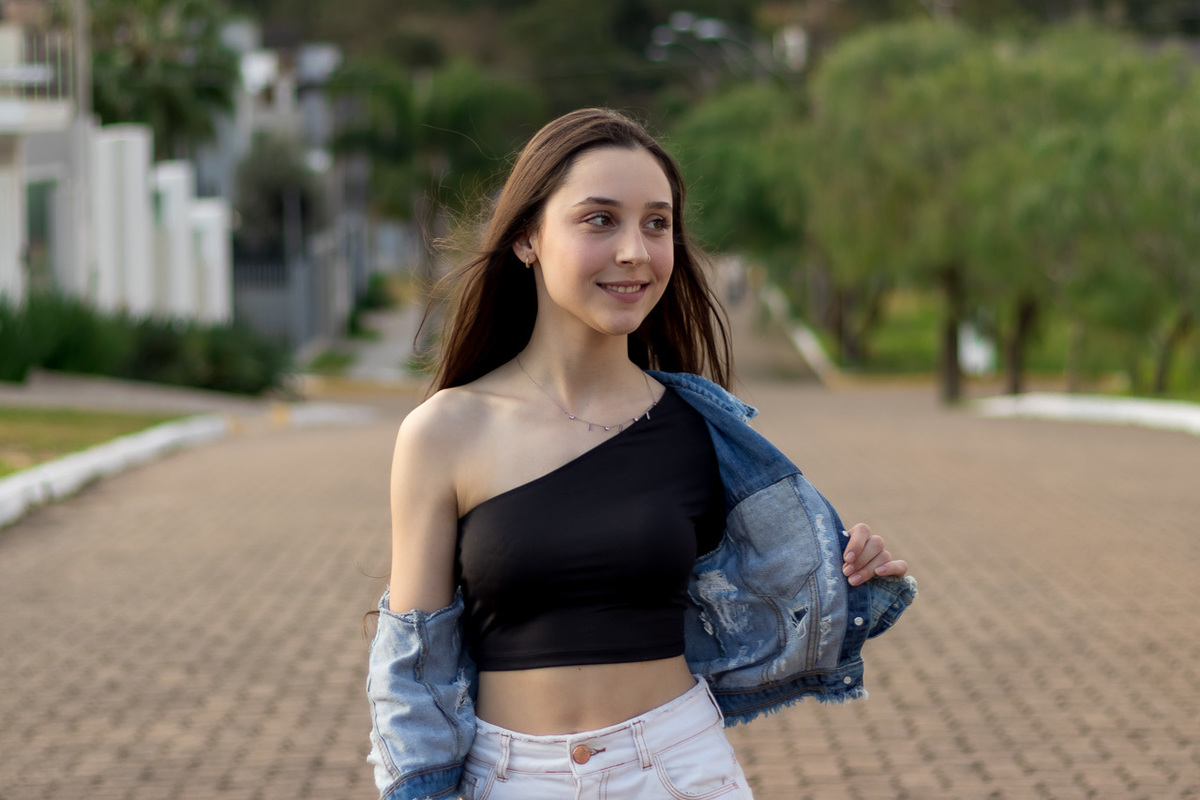 modelo caminha em direção da câmera no meio da rua. usa blusa preta com um ombro de fora e barriga também exposta. calça branca e uma jaqueta jeans caída nas costas