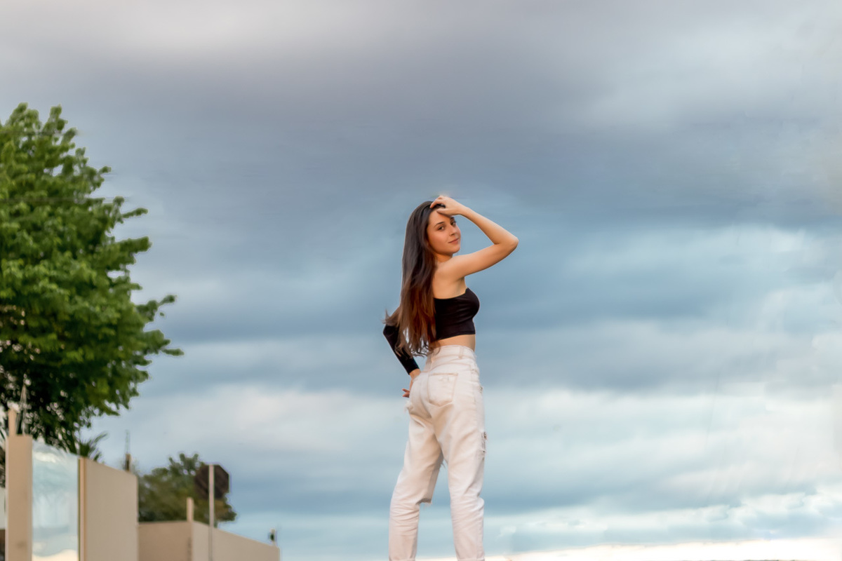 modelo de costas virando a cabeça para a câmera com a mão na cabeça. calça branca e blusa com um ombro de fora