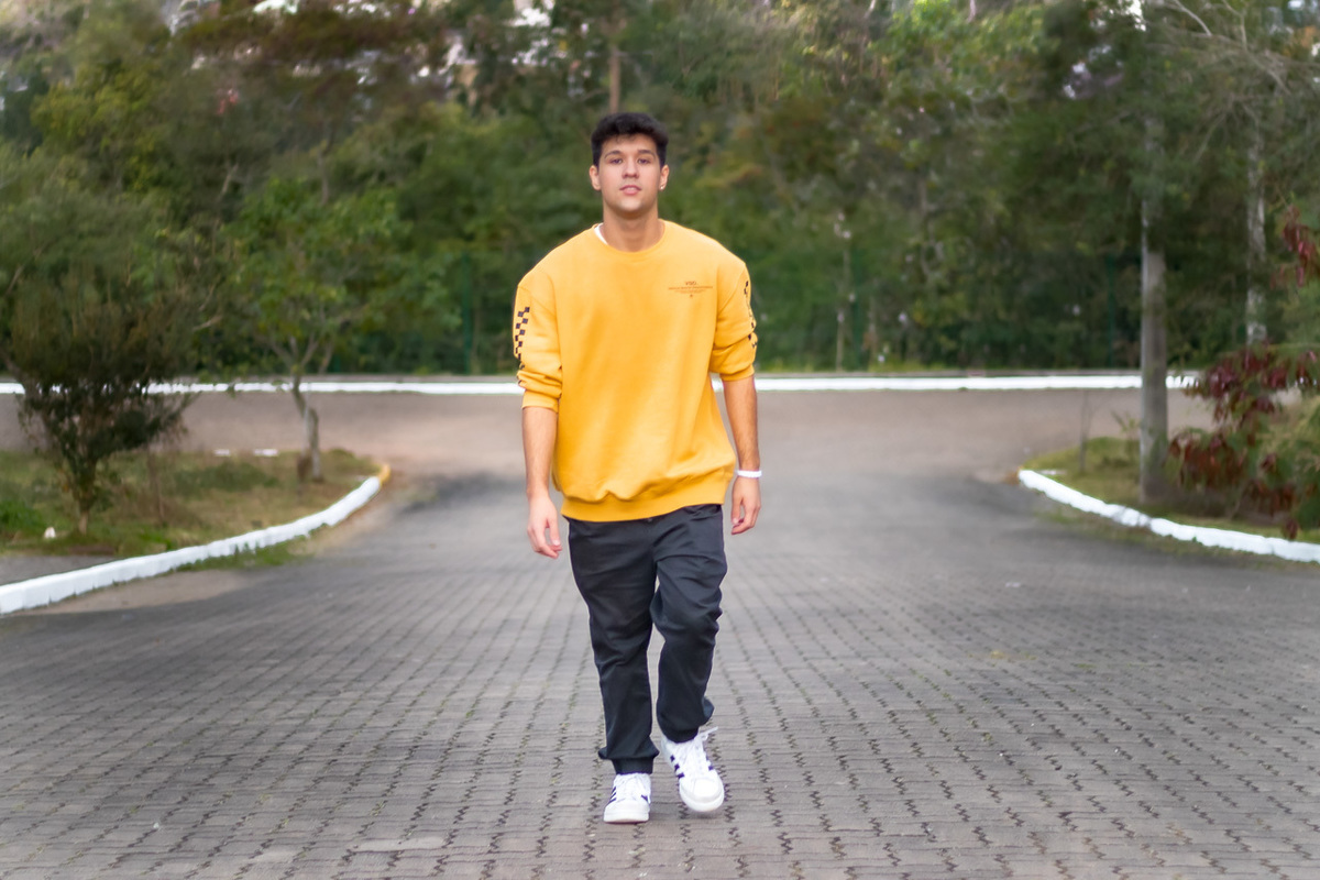 garoto de calça jeans tenis branco e moletom amarelo. Caminhando no meio de uma rua de calçamento em direção à câmera.