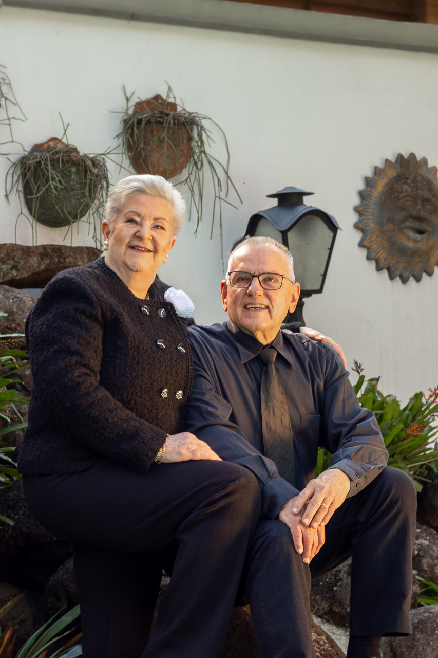 casal de terceira idade olhando para a câmera ela com o braço por tras dele apoiando a mão no ombro . Ambos cabelos grisalhos sorrindo. 