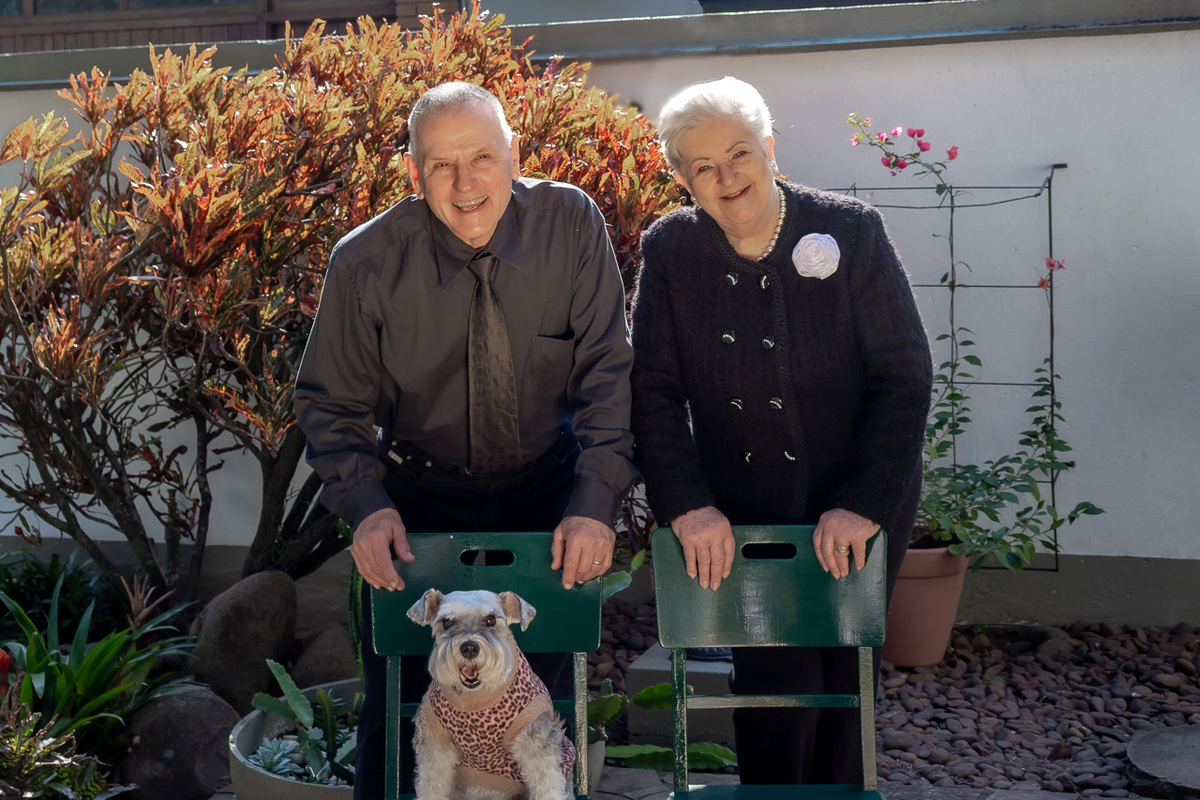 casal de cabelos grisalhos apoiados em duas cadeiras verdes. um cão schnauzer em cima de uma das cadeiras. no fundo parte do jardim
