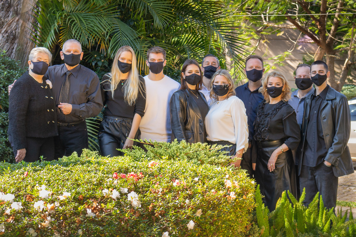 foto de família , 11 onze pessoas em pé lado a lado atrás de um arbusto de flores todos de máscara.