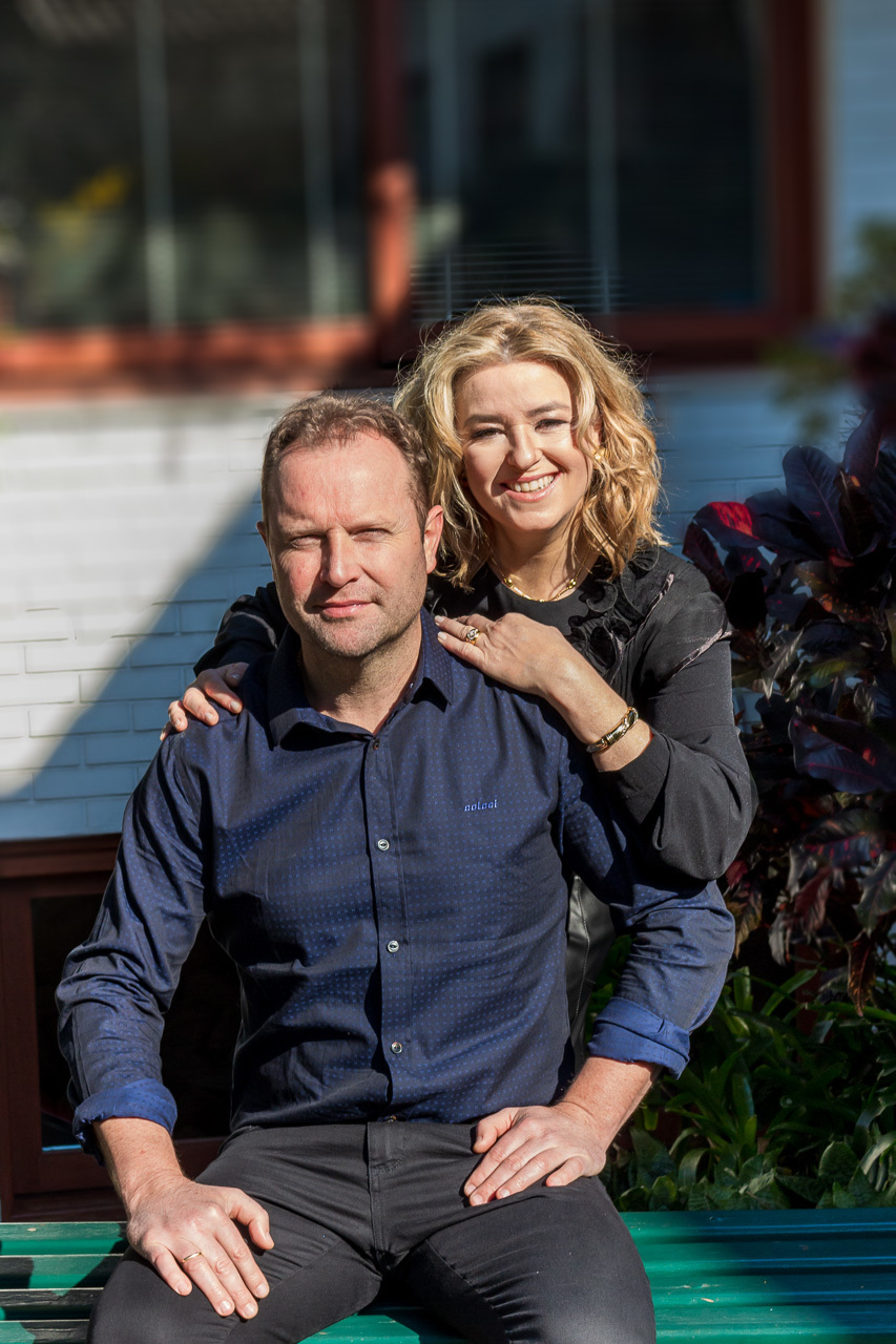 casal ele sentado em um banco ela atrás mais para o lado esquerdo com as mãos nos ombros dele. ela loira ondulado sorrindo ele cabelo curto cresppo castanho