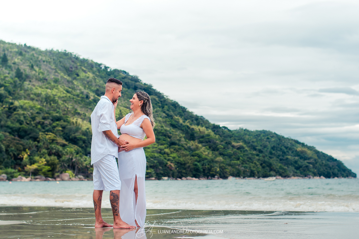 mulher grávida abraçada com o marido e sorrindo em ensaio gestante na praia em ubatuba são paulo