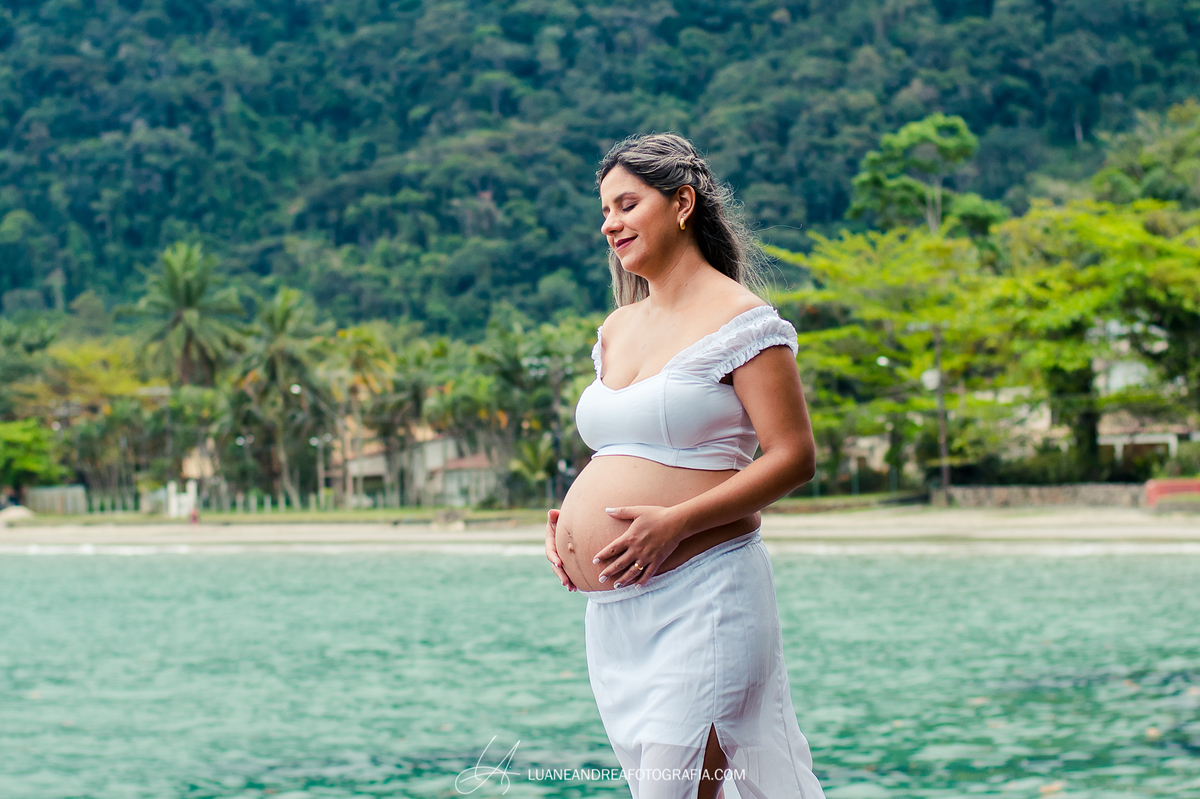 mulher grávida segurando a barriga e sorrindo na praia em ensaio gestante em ubatuba são paulo