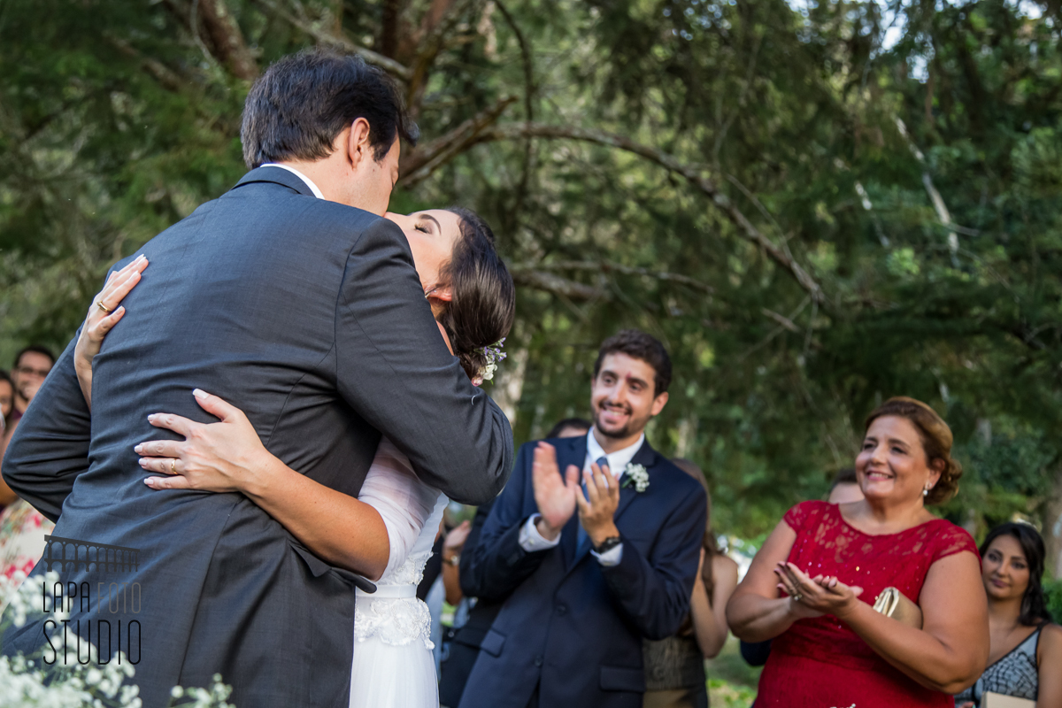 Beijo do casal durante a cerimônia de casamento com pais aplaudindo ao fundo.