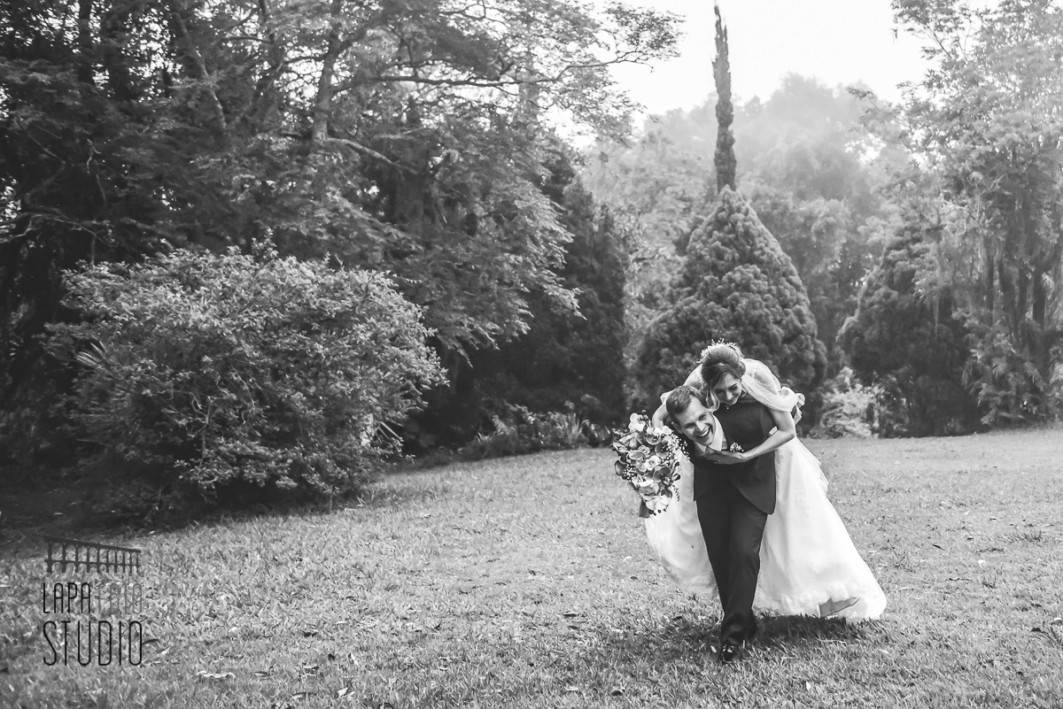 Foto em preto e branco do noivo carregando a noiva nas costas no gramado da Mansão das Heras