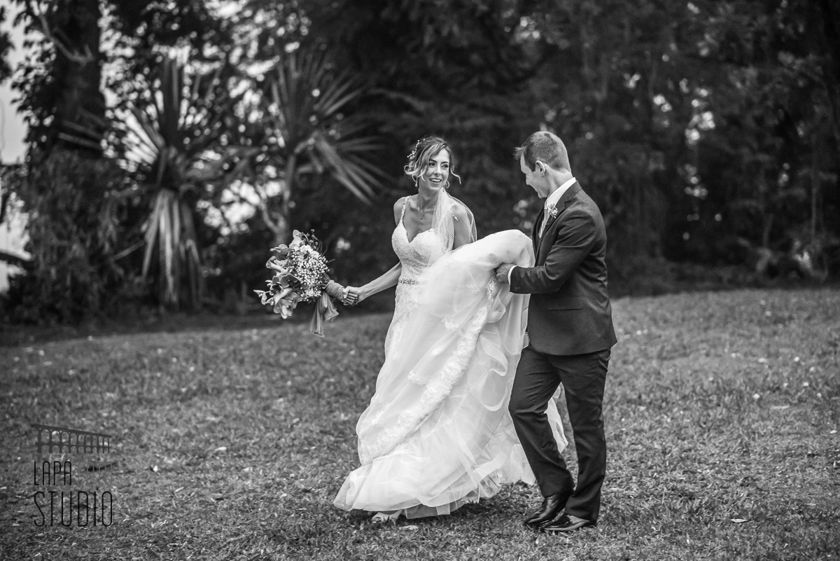 Foto em preto e branco do noivo ajudando a noiva a carregar seu vestido de casamento no jardim da Mansão das Heras