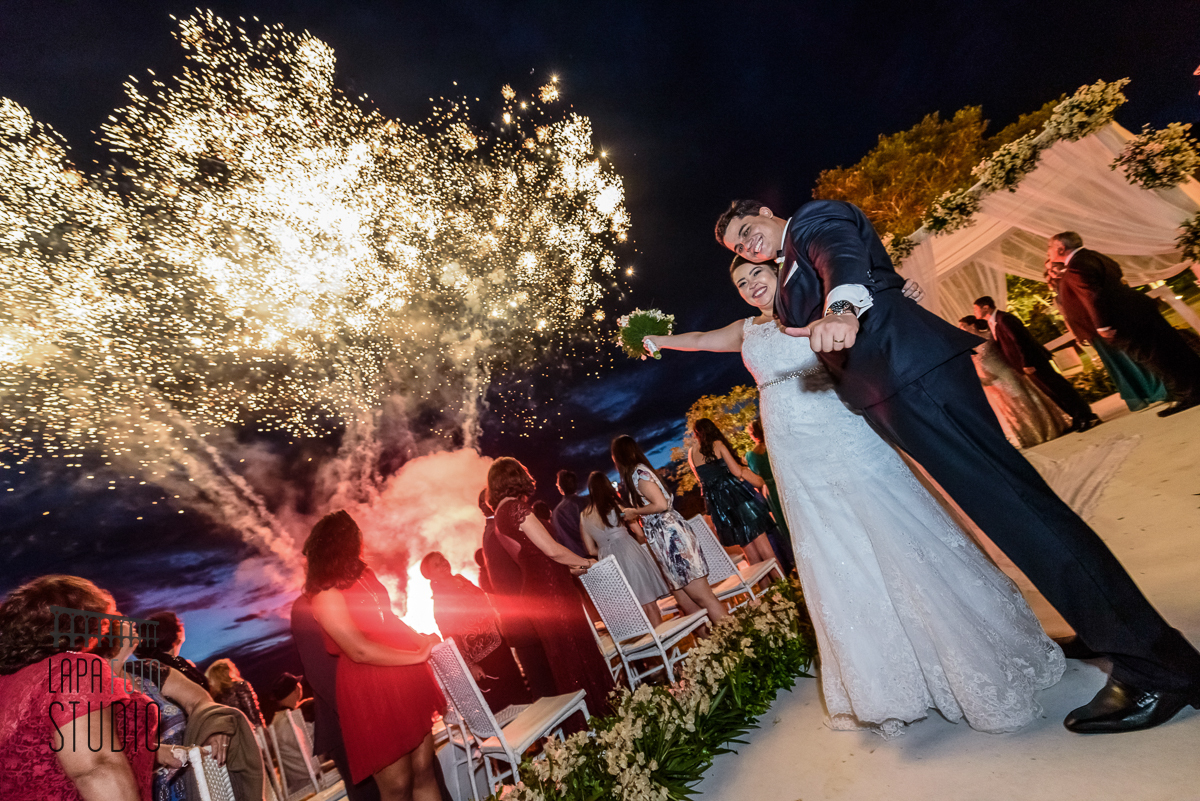 Noivos sorriem para a foto com queima de fogos ao fundo após a cerimônia