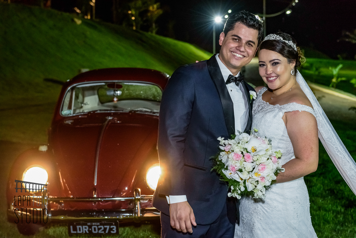 Noivos sorriem com fusca vermelho ao fundo 