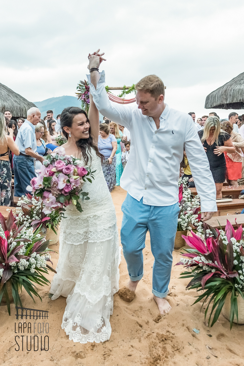 Noivos comemoram de braços para o alto na saída da cerimonia de casamento na praia