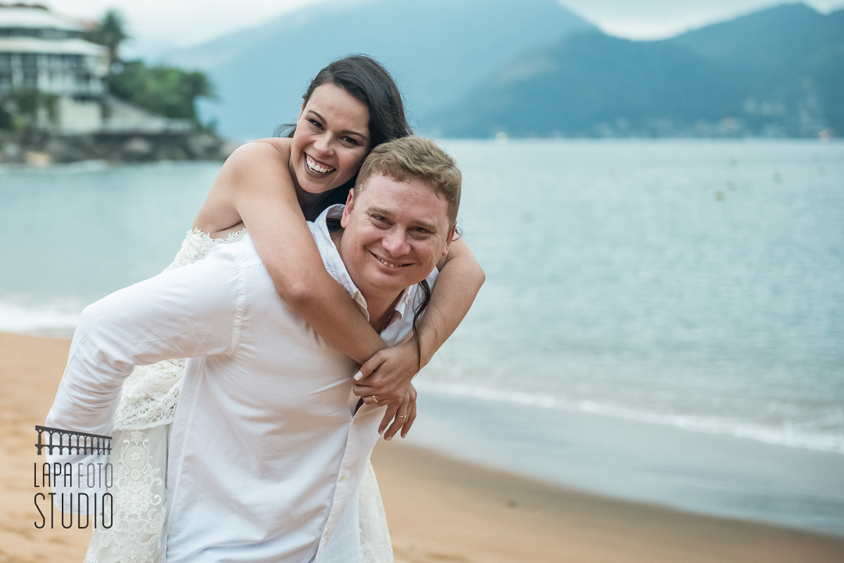 Noivo carrega a noiva nas cotas e os dois sorriem durante ensaio na praia