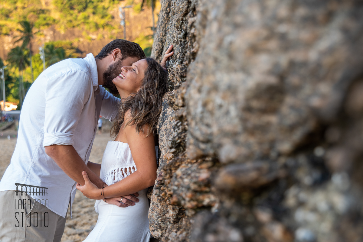 Noivo da um cheiro na noiva e ela sorri de olhos fechados no canto esquerdo da praia vermelha