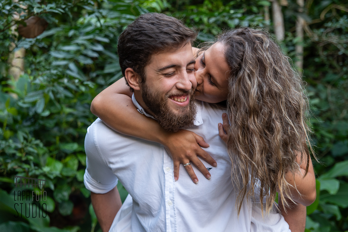 Noiva se debruça nas costas do noivo e dá um beijo nele no dia do ensaio pré casamento