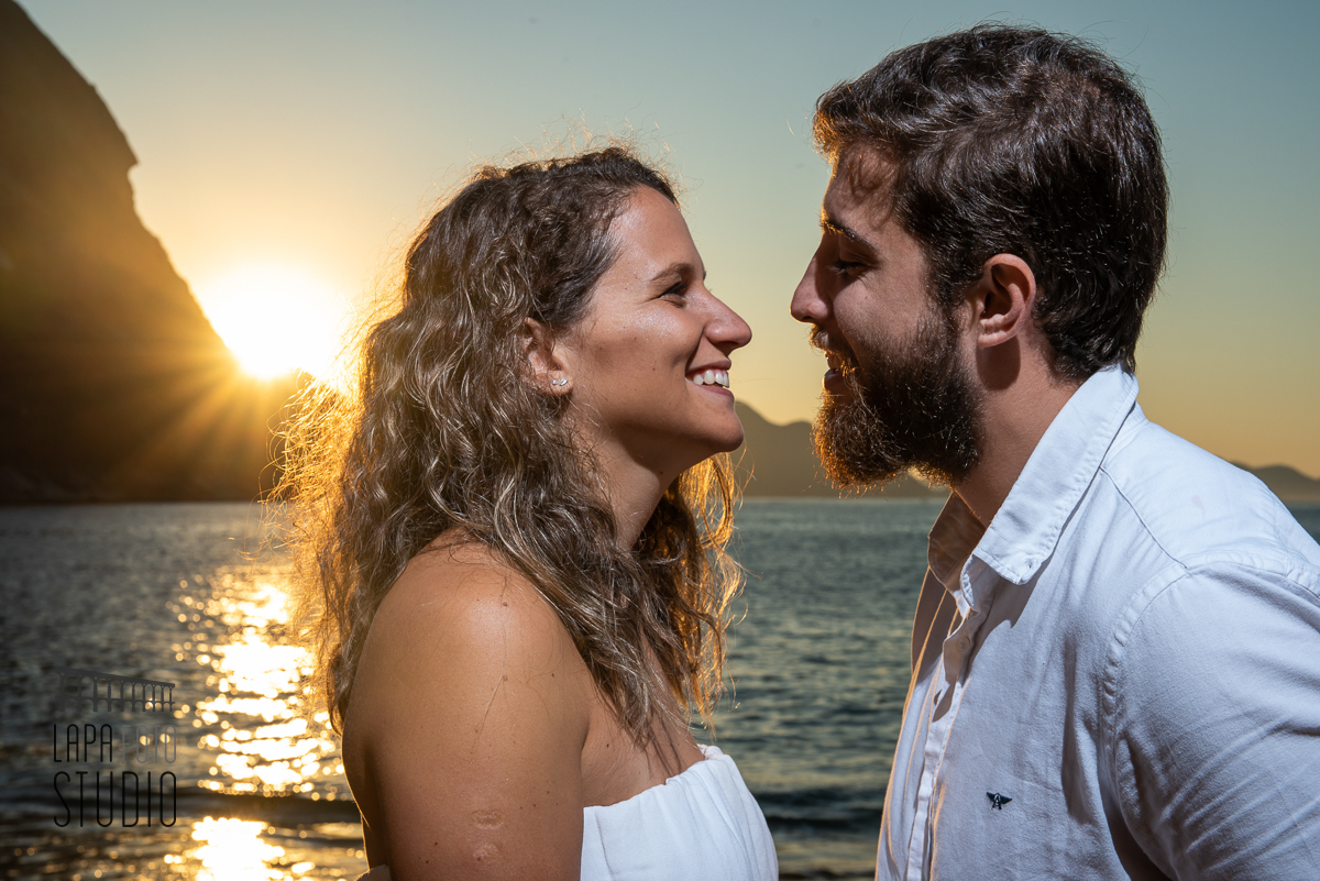 Noivos sorriem com mar ao fundo e sol nascendo na praia vermelha