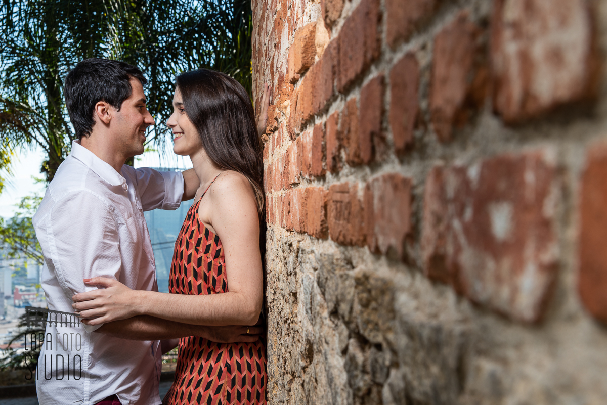 Noivos trocam olhar encostados na parede de tijolinhos no pré wedding no Parque das Ruínas