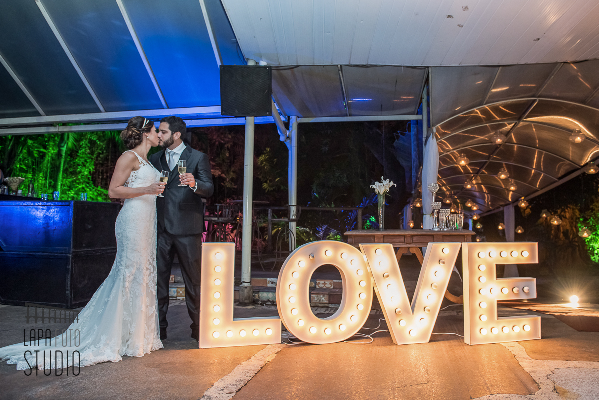 Noivos se beijando ao lado da palavra love iluminada na Casa do Flamboyant