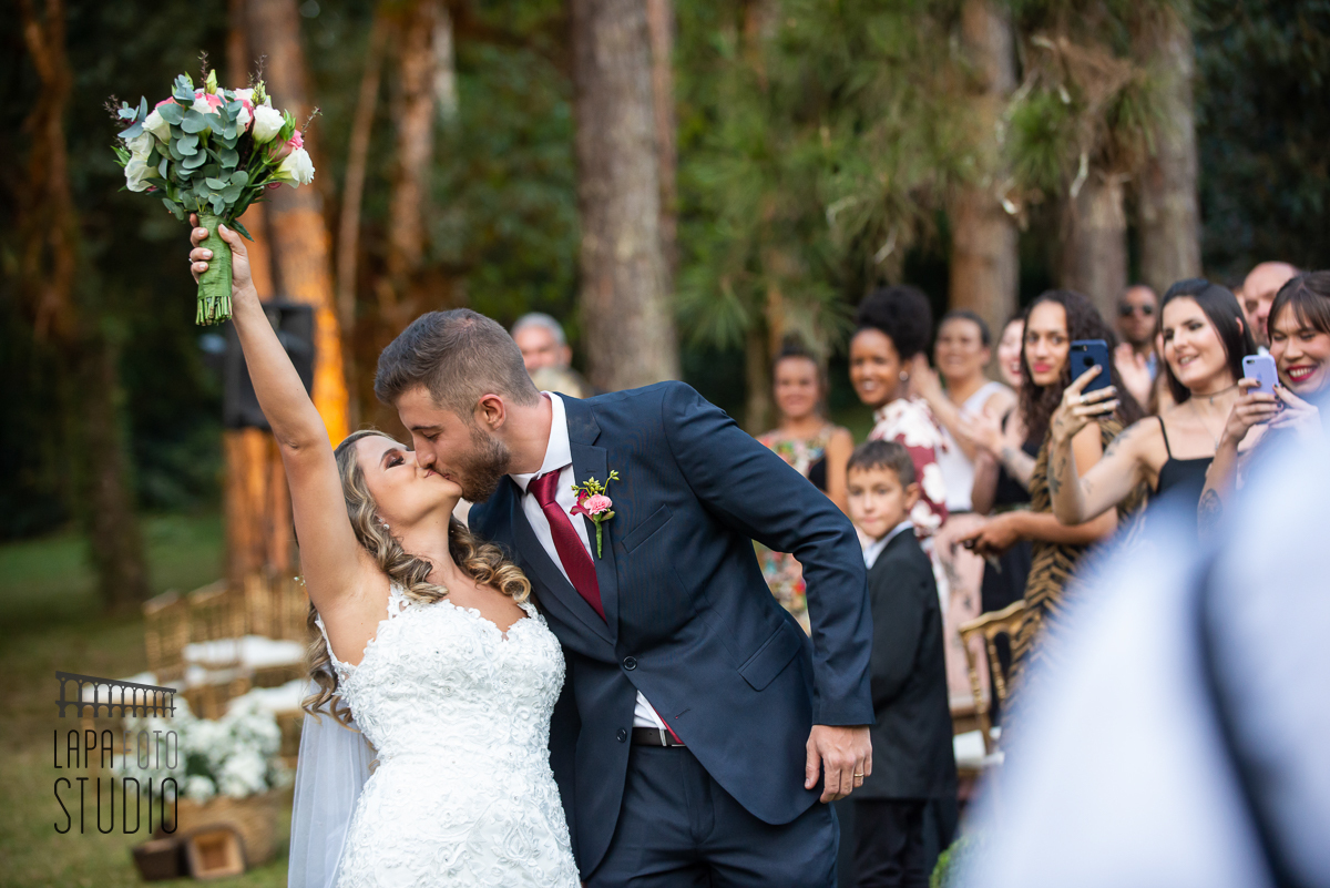 Noiva levanta o buquê enquanto o noivo a beija ao final da cerimônia de casamento