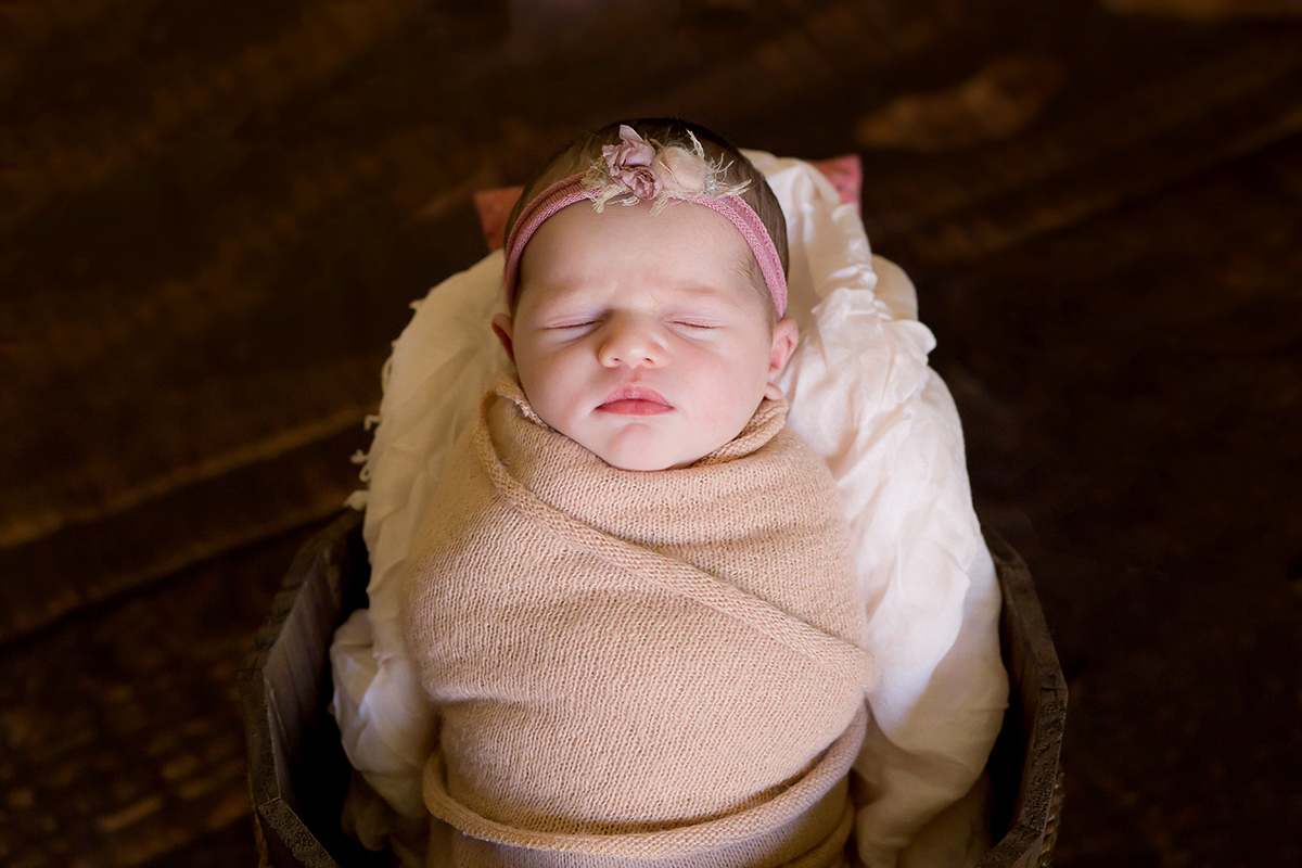 Fotografia de newborn em Pará de Minas pose no prop. Bebê enrolado no wrap. Newborn usando tons de rosa no piso de madeira.