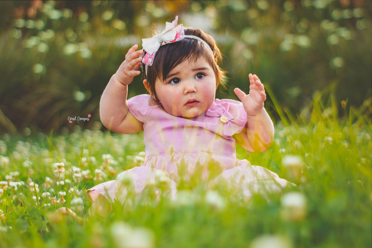 Fotografia de menina de 1 aninho sentada no chão no meio de flores com a mão na cabeça em ensaio realizado em Caçapava do Sul, RS