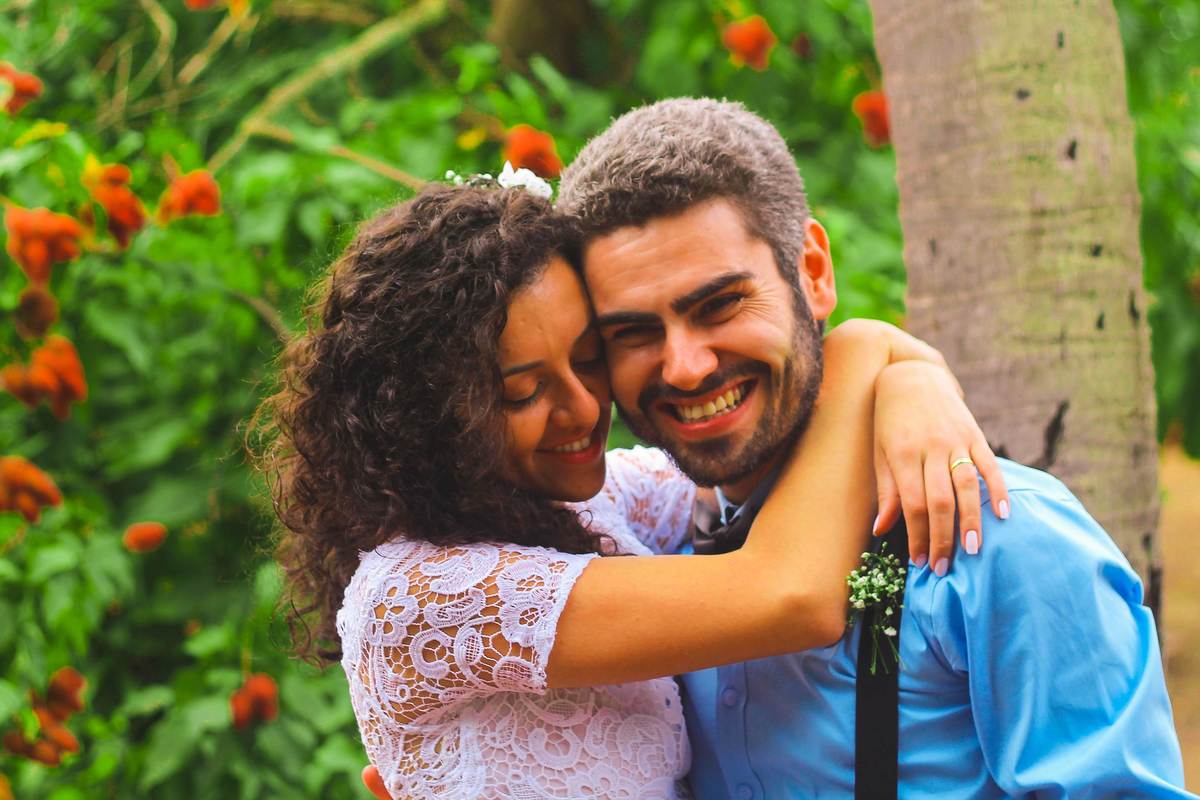 Fotografia dos noivos sorrindo abraçados próximo a árvore de urucu durante ensaio fotográfico pós casamento realizado na cidade de Jampruca Minas Gerais 