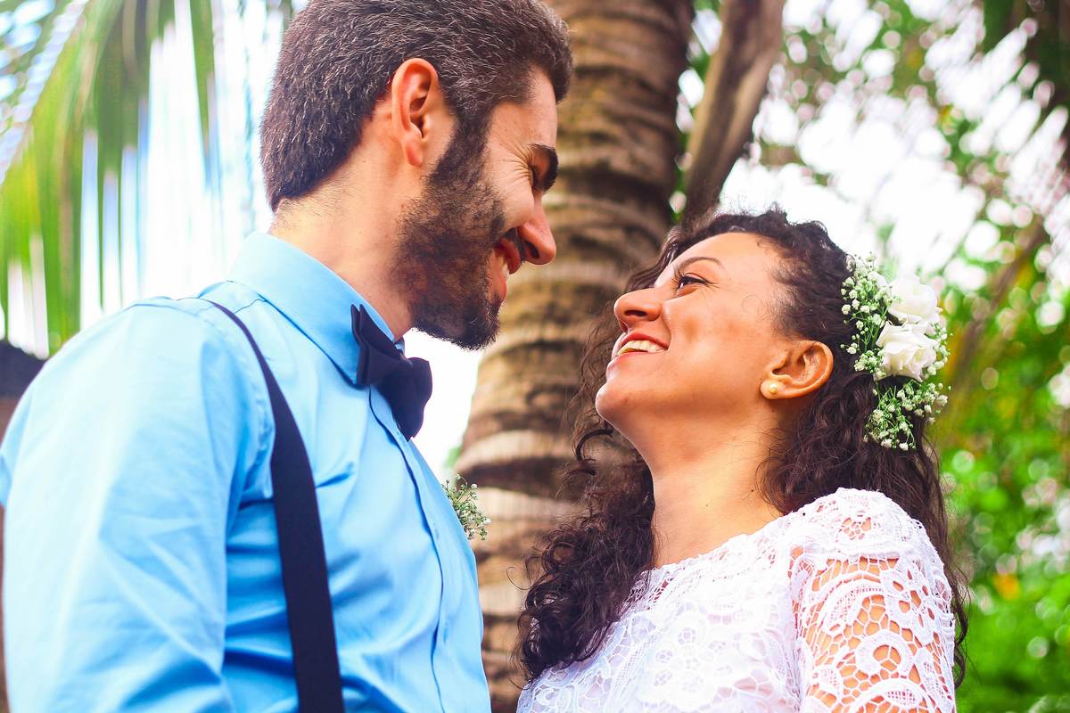 Fotografia dos noivos sorrindo e se olhando, próximo a coqueiro durante ensaio fotográfico pós casamento realizado na cidade de Jampruca Minas Gerais 