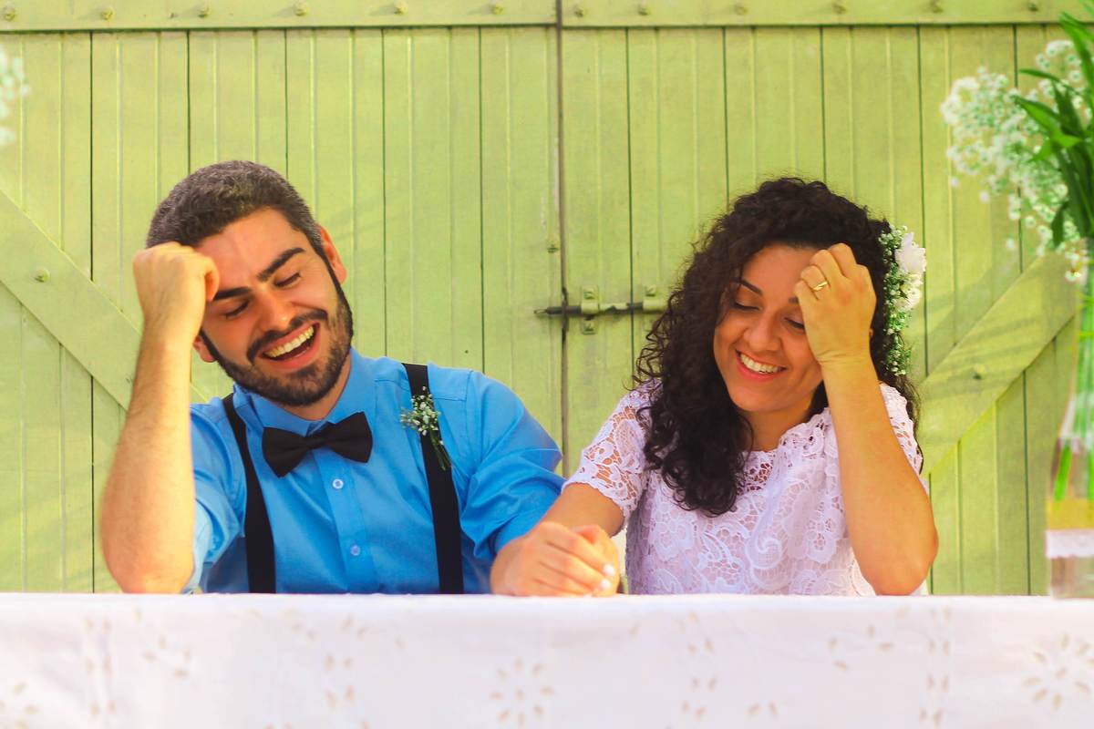 Fotografia dos noivos sorrindo com as mãos no cabelo próximo a portão de madeira e vasos flores durante ensaio fotográfico pós casamento realizado na cidade de Jampruca Minas Gerais 