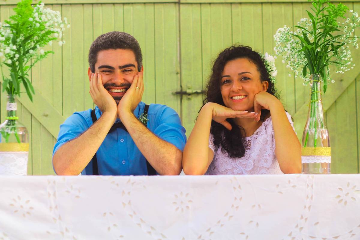 Fotografia dos noivos sorrindo posando com as mãos no rosto próximo a portão de madeira e vasos flores durante ensaio fotográfico pós casamento realizado na cidade de Jampruca Minas Gerais 