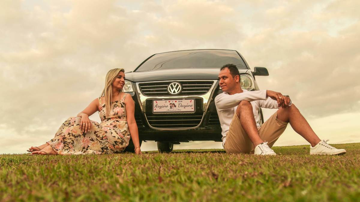 Foto dos noivos sentados na grama se olhando, em frente a carro, em  ensaio fotográfico pré casamento feito em Nova Lima - MG
