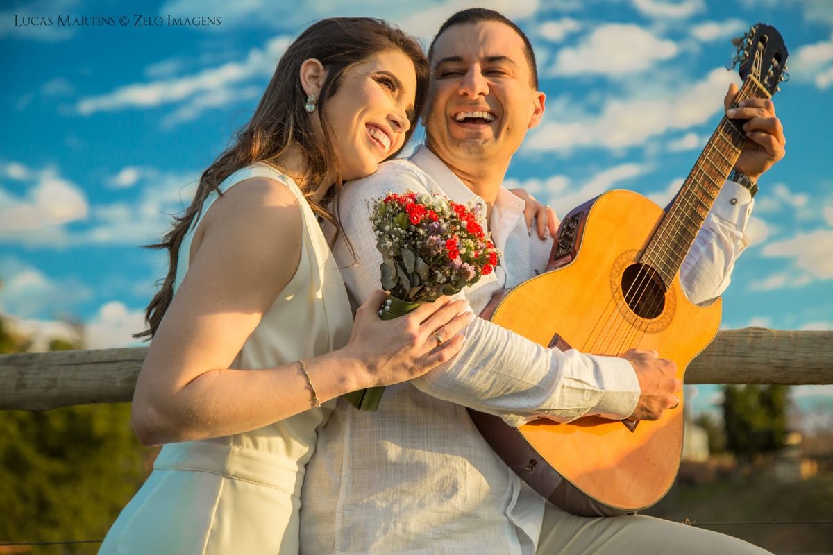 casal sorrindo no céu azul aberto enquanto o noivo toca violão