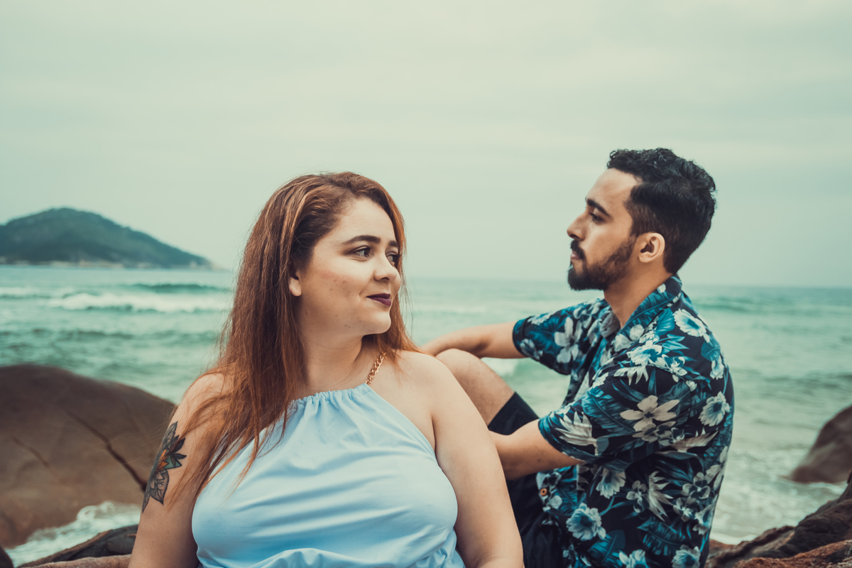 fotografia do casal, rapaz com blusa azul com estampa florida sentado numa pedra olhando em direção da namorada que usa um vestido  branco apoiada na pedra que o namorado dela está sentado, olhando na direção dele.