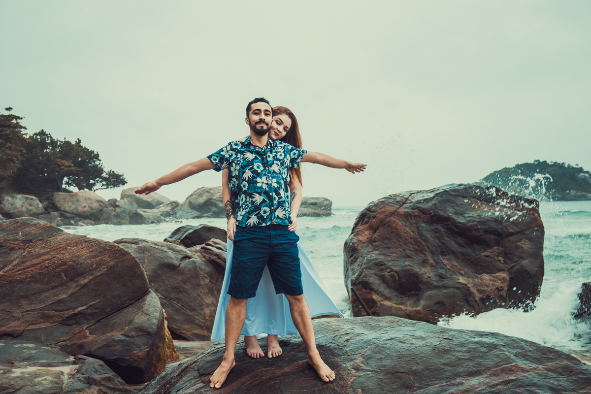 fotografia do casal em cima das pedras próximo ao mar, namorado está de braços abertos em frente a sua namorada, como se estivessem no titanic, durante ensaio fotográfico de casal feito na praia de grumari, Rio de Janeiro - RJ