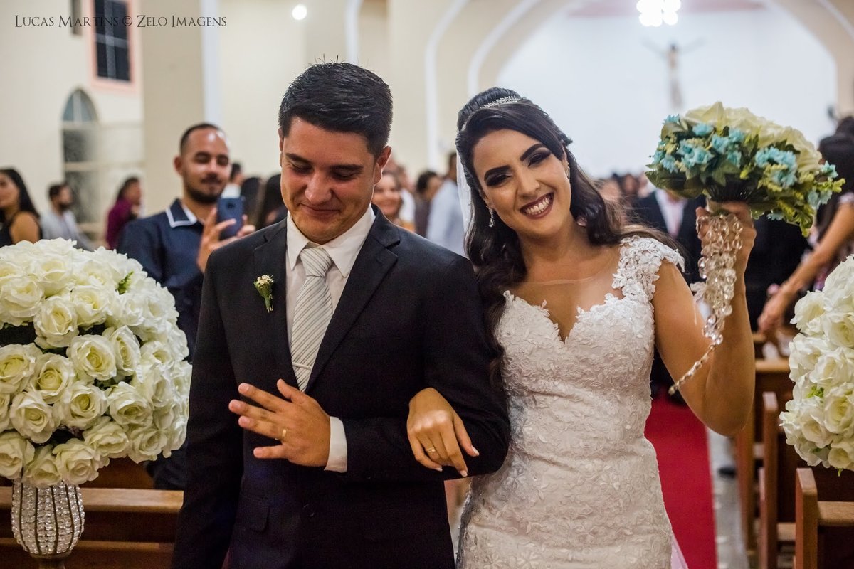 Fotografia da saída dos noivos, noiva erguendo o buquê e sorrindo durante casamento realizado na cidade de Virgem da Lapa - MG