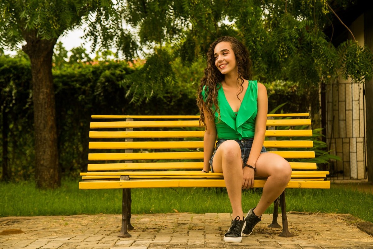 Debutante sorrindo sentada em um banco de madeira amarelo em ensaio 15 anos realizado na cidade de Araçuai, Minas Gerais