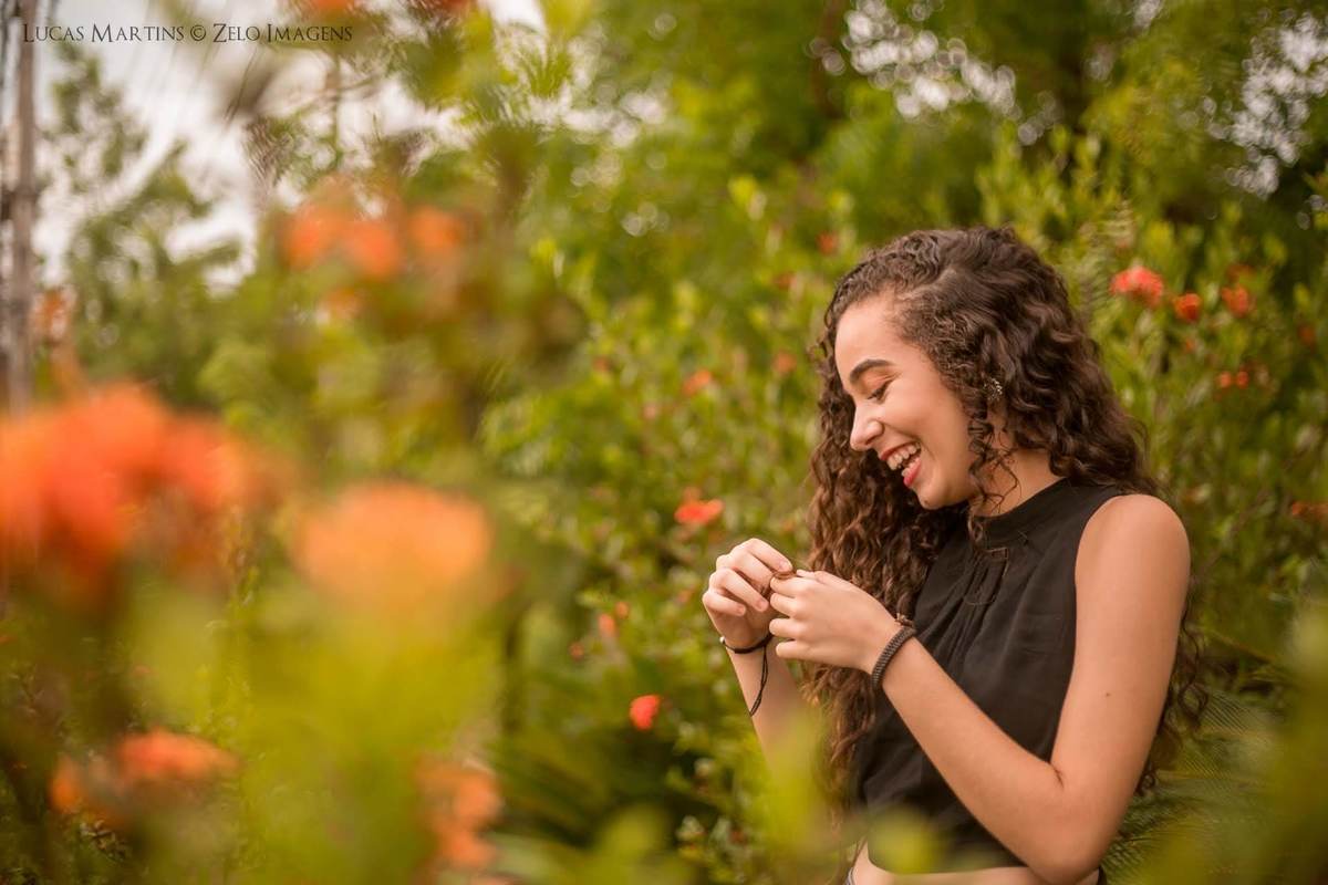Debutante sorrindo mexendo no cabelo em um campo de flores em ensaio 15 anos feito na cidade de Araçuai, Minas Gerais